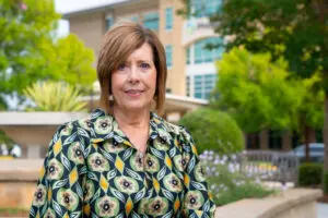 Ann Beaver stands outside in a patterned blouse, with greenery and a building in the background—A Place Close to Home.