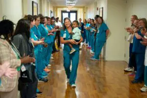 A nurse carrying a child walks down a hallway during the Sumter Pink Walk as colleagues in scrubs applaud on both sides.