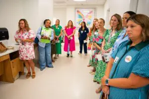 New NICU Mural Will Help Families Celebrate Their Babies' Journey Home 5 Women in colorful dresses stand in a bright hallway by a NICU mural featuring a butterfly painting in the background.