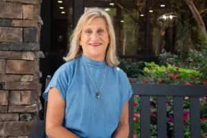 Smiling woman in a blue top sits on a bench outdoors by flowers and a stone wall, radiating hope and joy.