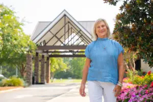 Smiling woman in a blue shirt stands outside near flowers on a sunny day, radiating hope.