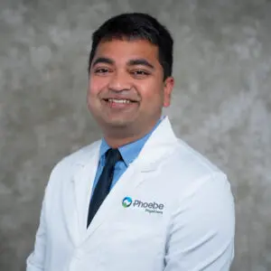 Smiling man in a white doctor’s coat and blue shirt with tie, standing in front of a gray mottled background.
