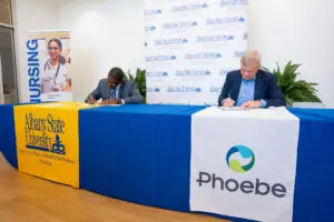 Two men in business attire sign a document at a table featuring Albany State University and the Phoebe nursing program branding.