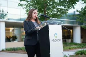 A woman in a black blazer speaks at a lectern branded with the Phoebe organization logo at a Wave of Light remembrance event.