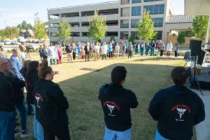 A large group of emergency medical staff from Phoebe in scrubs and hoodies gather around a speaker on a lawn at an event.