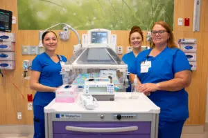 Three nurses in blue scrubs stand behind a medical cart and infant incubator during nursing education at a hospital.