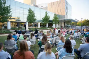 A speaker addresses a large outdoor audience seated in white chairs before a glass building for a Pregnancy and Infant Loss vigil.