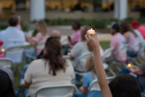 A hand holds a lit candle for a Wave of Light remembrance above a crowd seated in outdoor chairs.