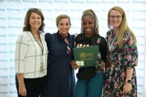 Four women stand before a backdrop for the organization Phoebe Sumter, one holding a DAISY Award certificate and sculpture.