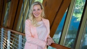 A woman with long blonde hair in a pink blazer and white top stands by a railing and windows, smiling for a professional bio photo.