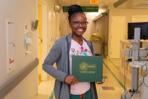 A smiling PPMH Labor & Delivery Nurse stands in a hallway holding a green “DAISY Award” certificate, wearing glasses, a gray hoodie, colorful lanyard, and hospital ID badge. The background shows medical equipment and doors.