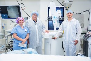 Three medical professionals in scrubs and protective gear stand in a hospital room with advanced equipment, smiling at the camera.