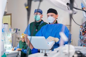 Several medical professionals in scrubs and masks operate advanced equipment in a bright hospital room, focusing on a monitor.