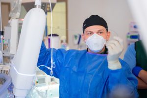 A medical professional in a blue gown, black cap, and face mask handles a thin wire during a lung procedure in a hospital.