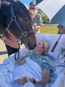 A hospice patient in a hospital bed outdoors lovingly touches noses with her horse as people nearby hold an umbrella to shade her.
