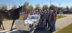 A group stands outside by a hospice patient in a hospital bed, with a saddled horse nearby; final goodbyes in the sun.
