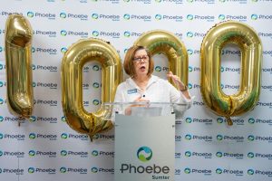 A woman speaks at a podium featuring the Phoebe Sumter logo, celebrating 1,000 robotic procedures amid gold "1000" balloons.