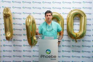 A man in scrubs stands at a Phoebe Sumter podium, flanked by gold "1,000" balloons, celebrating robotic procedure milestones.