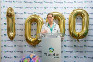 A woman stands at a Phoebe Sumter podium, speaking before a backdrop with Phoebe logos and gold 1000 balloons celebrating a milestone.