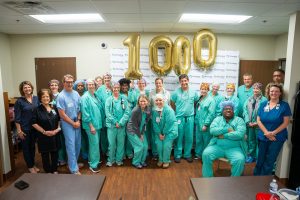 A group of healthcare workers in green scrubs smile together with gold 1000 balloons and a Phoebe Sumter backdrop celebrating 1,000 robotic procedures.