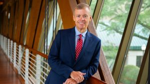 A man in a navy blue suit and red striped tie smiles in a modern glass hallway, symbolizing fair employee compensation practices.