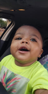 A smiling toddler with short hair in a bright yellow shirt sits in a car seat; mom credits Phoebe Worth ER with saving her baby’s life.