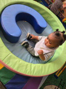 A baby with two pigtails sits on a round mat, looking curious—her expression is worthy of a dramatic ER scene.