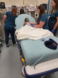 A baby lies on a hospital bed in the Phoebe Worth ER as three healthcare workers in blue scrubs provide care. Medical equipment is visible.