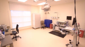 Bright, clean AAPHC medical exam room with chairs, equipment, cabinets, monitors, and window. Space supports colon cancer awareness.