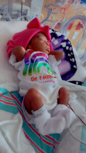 A premature baby in a NICU incubator wears a rainbow onesie, pink bow hat, hospital bands, and soft blankets—symbolizing hope.