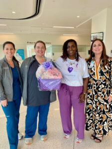 Four women smile together in the NICU, one holding a purple gift basket. They wear scrubs, a printed dress, and casual clothes.