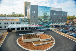 A modern Phoebe Hospitals building with large glass windows, Emergency & Trauma Center sign, cars parked, and landscaped entrance.