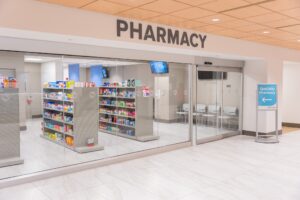 A modern pharmacy with glass walls, stocked shelves, a waiting area, ACHC-accredited sign, tiled floor, and bright lighting.