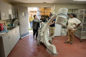 Four people move imaging equipment through a doorway as two others observe in a room with red floors and white cabinets.