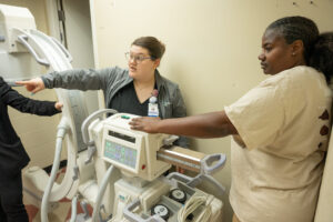 Two women use imaging equipment in a clinical setting, while a third person points off-camera, showcasing veterinary technology.