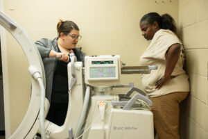 Two women stand by advanced imaging equipment in a beige room, one at the controls and one in tan scrubs observing attentively.