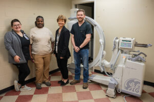 Four people smile beside advanced imaging equipment in a beige room with checkered tile floors and a large GE-branded machine.