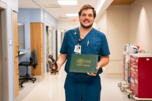 A nurse in blue scrubs stands in a hospital hallway, smiling and holding a green DAISY Award folder and a small sculpture.