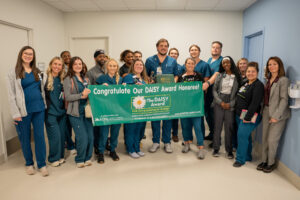 A group of compassionate Phoebe healthcare workers smile together in a hallway, holding a green DAISY Award Honoree banner.
