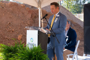 A man in a blue suit and yellow tie speaks at a podium with a Phoebe sign, under a tent. Ferns, another man, and dirt are visible.