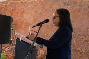 A woman wearing glasses and a navy suit speaks at a podium, with a dirt mound behind her—celebrating a new urgent care clinic in Lee County.