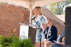 A woman in a white dress with blue flowers speaks at a Phoebe-logo podium at an urgent care event, two men seated behind her.