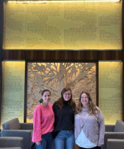 Three women smile standing before a decorative wall about pediatric organ donation, with tree design and beige armchairs nearby.