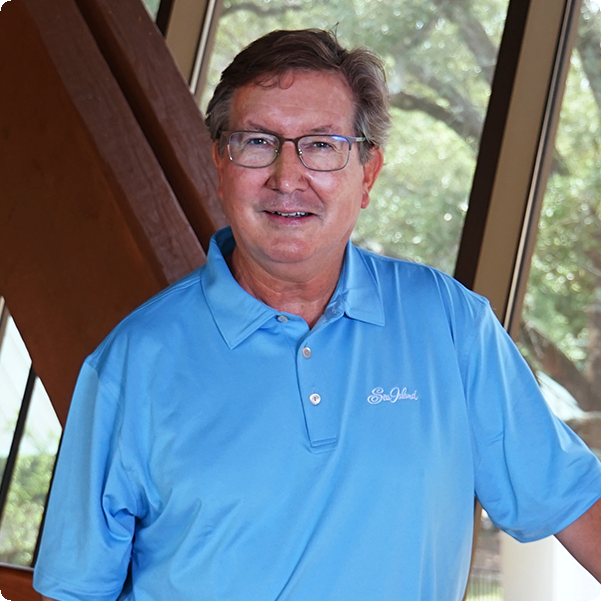 A smiling man in glasses and a light blue polo shirt stands indoors near large windows with trees visible outside.