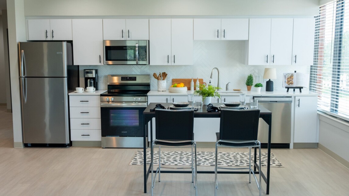 Modern kitchen in Living & Learning Housing with white cabinets, stainless steel appliances, a dining table set for four, and a window filling the space with natural light. Countertops display kitchen essentials, plants, and fresh fruit. Modern kitchen in Living & Learning Housing with white cabinets, stainless steel appliances, a dining table set for four, and a window filling the space with natural light. Countertops display kitchen essentials, plants, and fresh fruit.