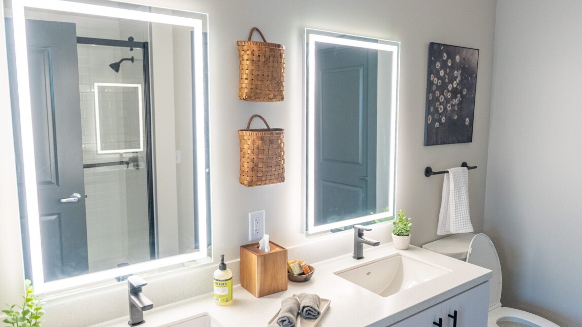 A modern bathroom in student housing features two sinks, illuminated mirrors, potted plants, a soap dispenser, wicker baskets on the wall, a towel rack, and glimpses of the shower and toilet—perfect for an educational community like Living & Learning Housing. A modern bathroom in student housing features two sinks, illuminated mirrors, potted plants, a soap dispenser, wicker baskets on the wall, a towel rack, and glimpses of the shower and toilet—perfect for an educational community like Living & Learning Housing.