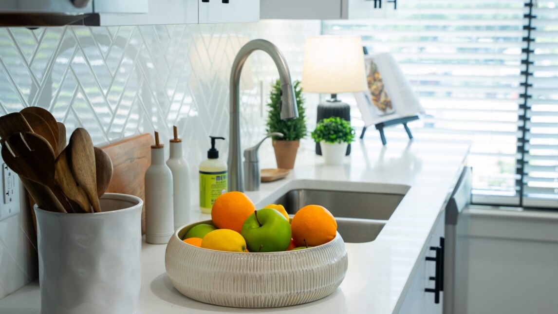 A modern kitchen countertop in Living and Learning Housing features a bowl of colorful fruit, wooden utensils, potted plants, a soap dispenser, and a lit lamp beside a stainless steel sink beneath a window with blinds—perfect for student housing. A modern kitchen countertop in Living and Learning Housing features a bowl of colorful fruit, wooden utensils, potted plants, a soap dispenser, and a lit lamp beside a stainless steel sink beneath a window with blinds—perfect for student housing.