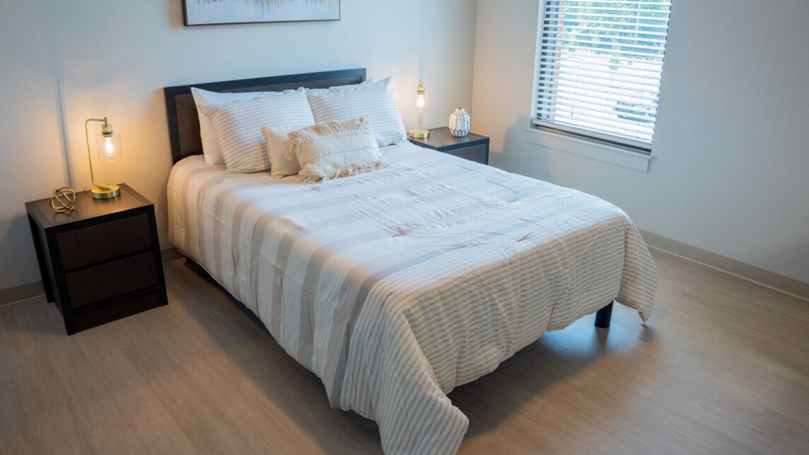A neatly made bed with white and beige striped bedding is centered between two dark nightstands with lamps in a bright student housing room with wood flooring, a large window with blinds, and a painting above the bed. A neatly made bed with white and beige striped bedding is centered between two dark nightstands with lamps in a bright student housing room with wood flooring, a large window with blinds, and a painting above the bed.