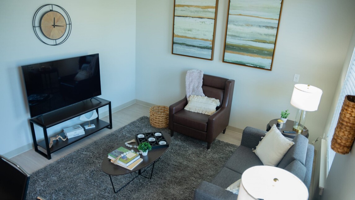 A modern Living & Learning Housing living room with a gray sofa, brown armchair, coffee table, flat-screen TV on a stand, two abstract paintings, wall clock, floor lamp, and textured gray rug. Natural light fills the space. A modern Living & Learning Housing living room with a gray sofa, brown armchair, coffee table, flat-screen TV on a stand, two abstract paintings, wall clock, floor lamp, and textured gray rug. Natural light fills the space.