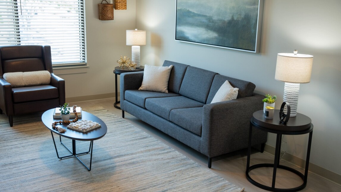A modern Living and Learning Housing living room with a gray sofa, armchair, two round side tables with lamps, and a coffee table on a light striped rug. A large painting hangs above the sofa, and window blinds let in natural light. A modern Living and Learning Housing living room with a gray sofa, armchair, two round side tables with lamps, and a coffee table on a light striped rug. A large painting hangs above the sofa, and window blinds let in natural light.